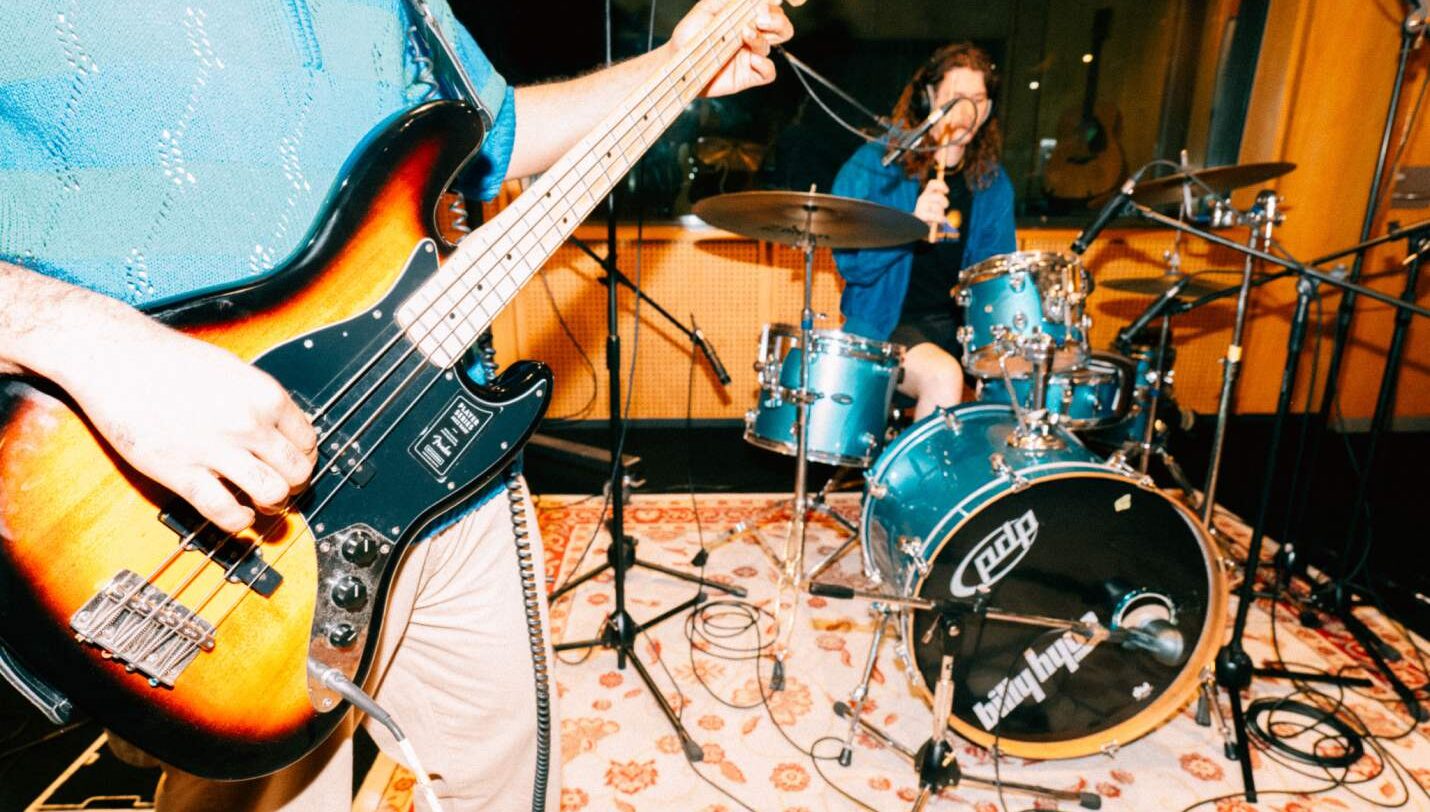 Two students in a recording studio together. One student playing an electric guitar and one students playing the drums | SAE Music