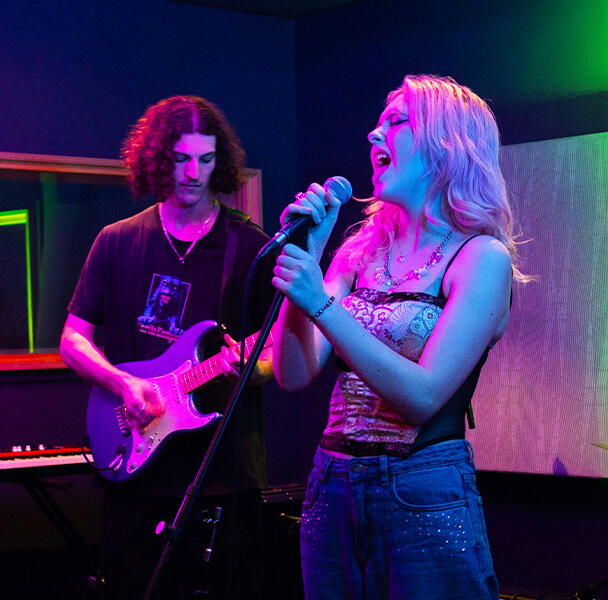 Singer sings into microphone. Musician stands behind with an electric guitar - playing. Both are in a recording studio with blue, green and pink lighting