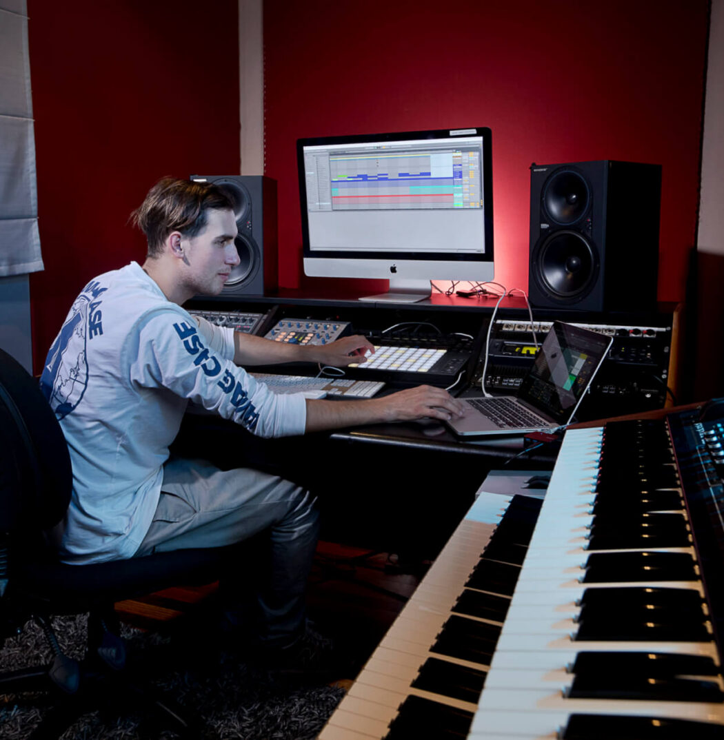 Man sits in an office chair with one hand on a Ableton Push and the other on laptop. He is in a music recording studio which includes a monitor with audio editing software, two speakers on top of desk, multiple mixing sound consoles and a tiered piano keyboard (two keyboards on top of each other). The keyboards have multiple sliders and dials.