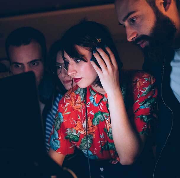 A group of people crowd around a laptop. The light from laptop lights up the faces of the people looking at it. A girl in the centre is the focal point. She is wearing headphones and also one hand on ear piece other on the desk in front.